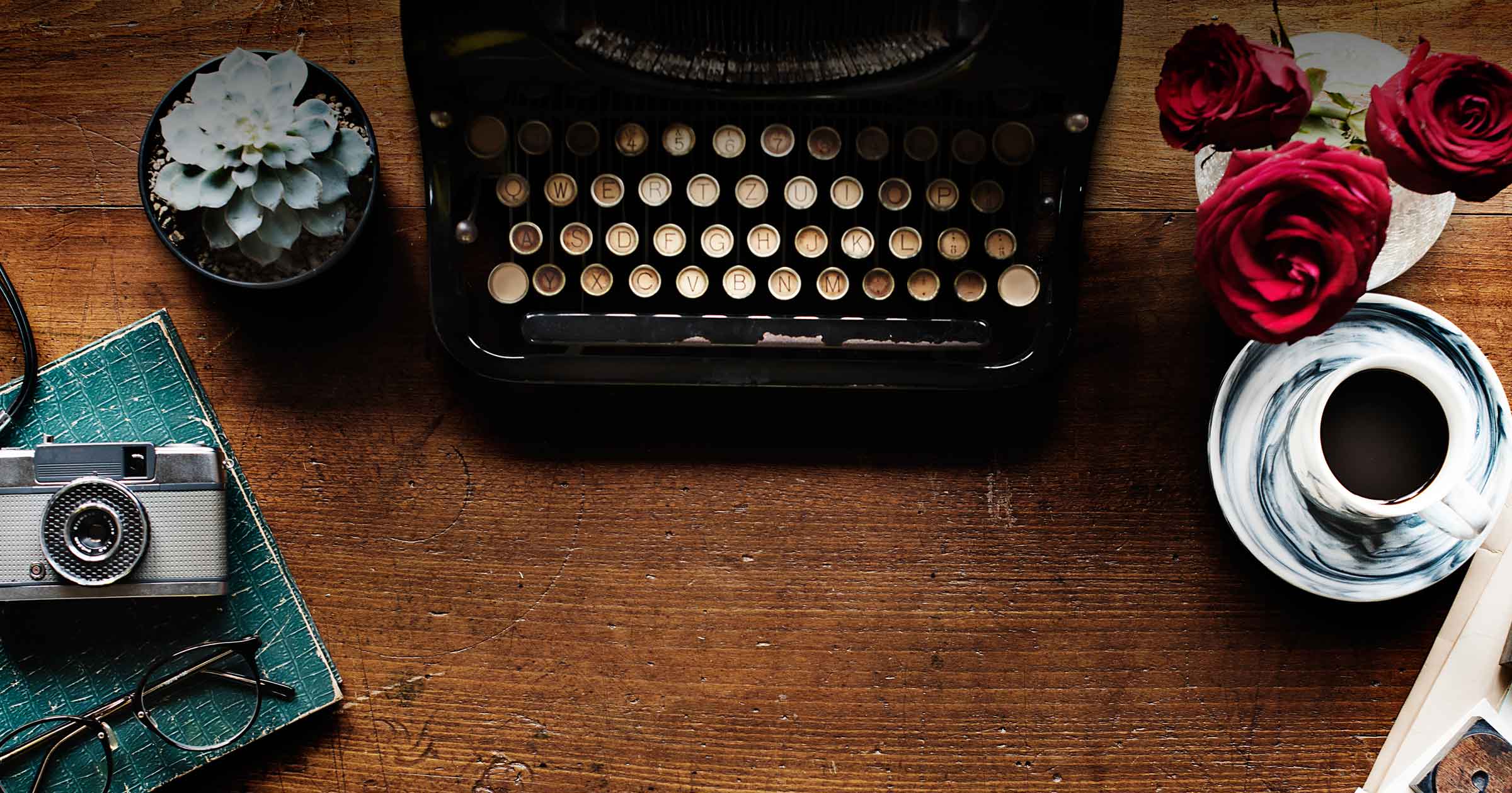 A typewriter sits on a desk, surrounded by other human interest artifacts, like coffee and a camera and flowers.