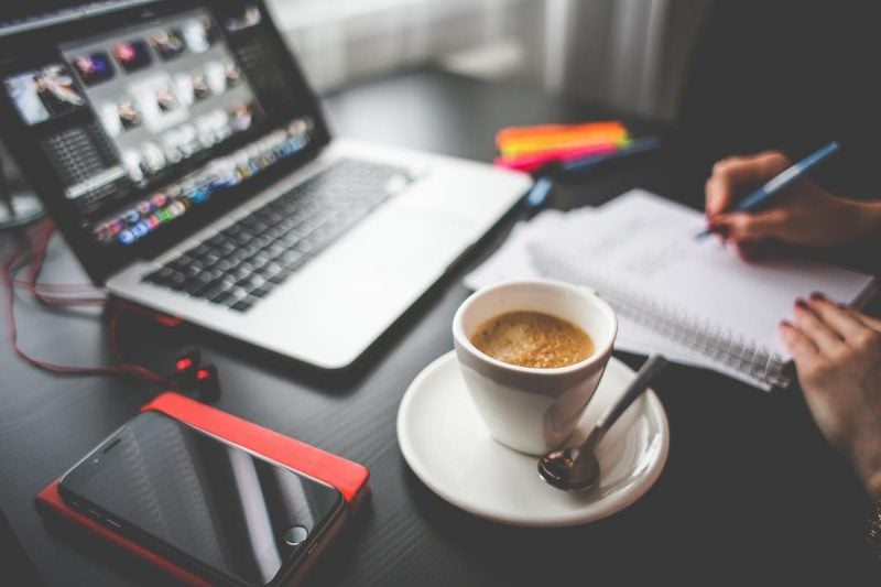 A laptop sits on a table in front of a person with a coffee, taking notes.