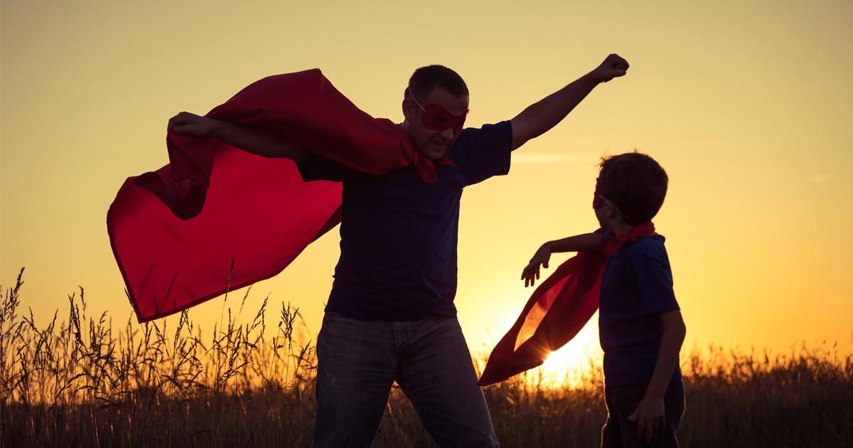 A father and son play in a field at sunset, wearing their capes and acting out superhero-hood!