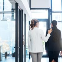 Two people walk down a company hallway and one person has their hand on their friend's shoulder.