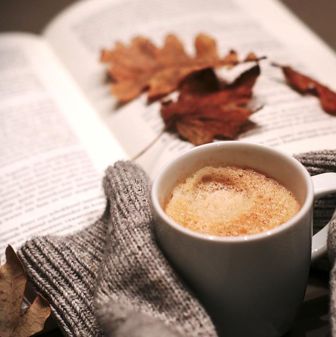 A latte rests beside a sweater and a book, maybe a journal, with some leaves on top.