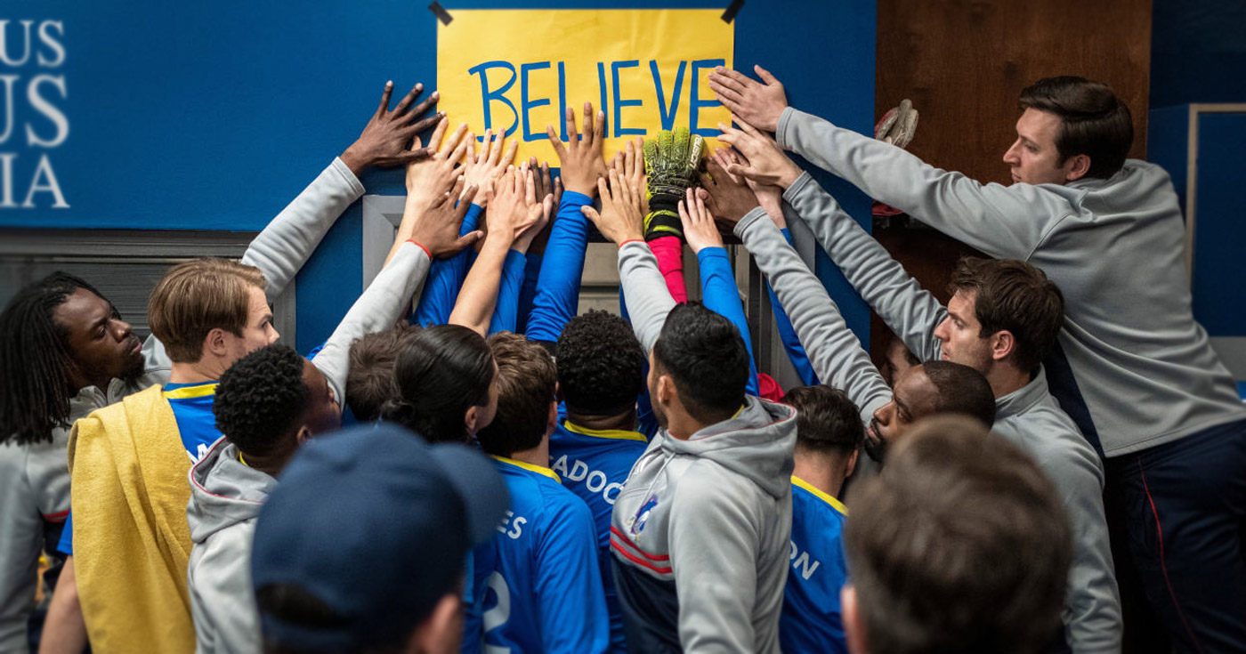Team members of AFC Richmond, from the Ted Lasso television series, stand under a beat up "BELIEVE" poster and touching it with their hands.