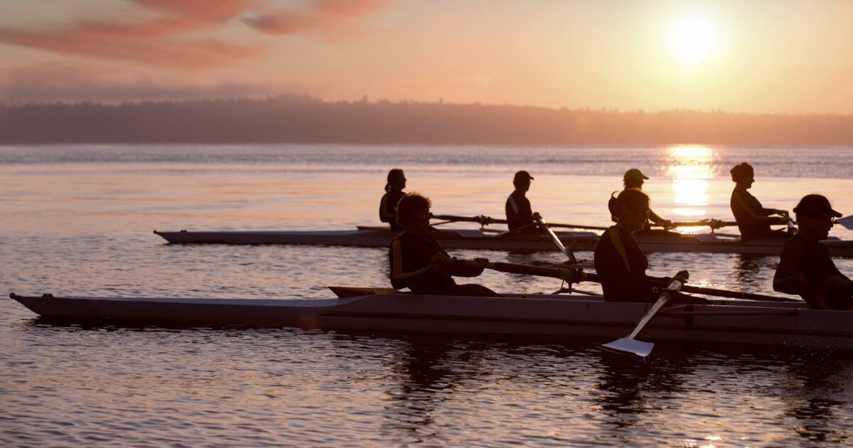 Two rowing teams take a rest on the water during practice and the sun rises in the background.